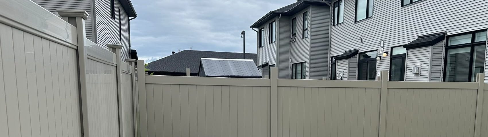 A long Adobe PVC privacy fence running between two rows of houses under an overcast sky.
