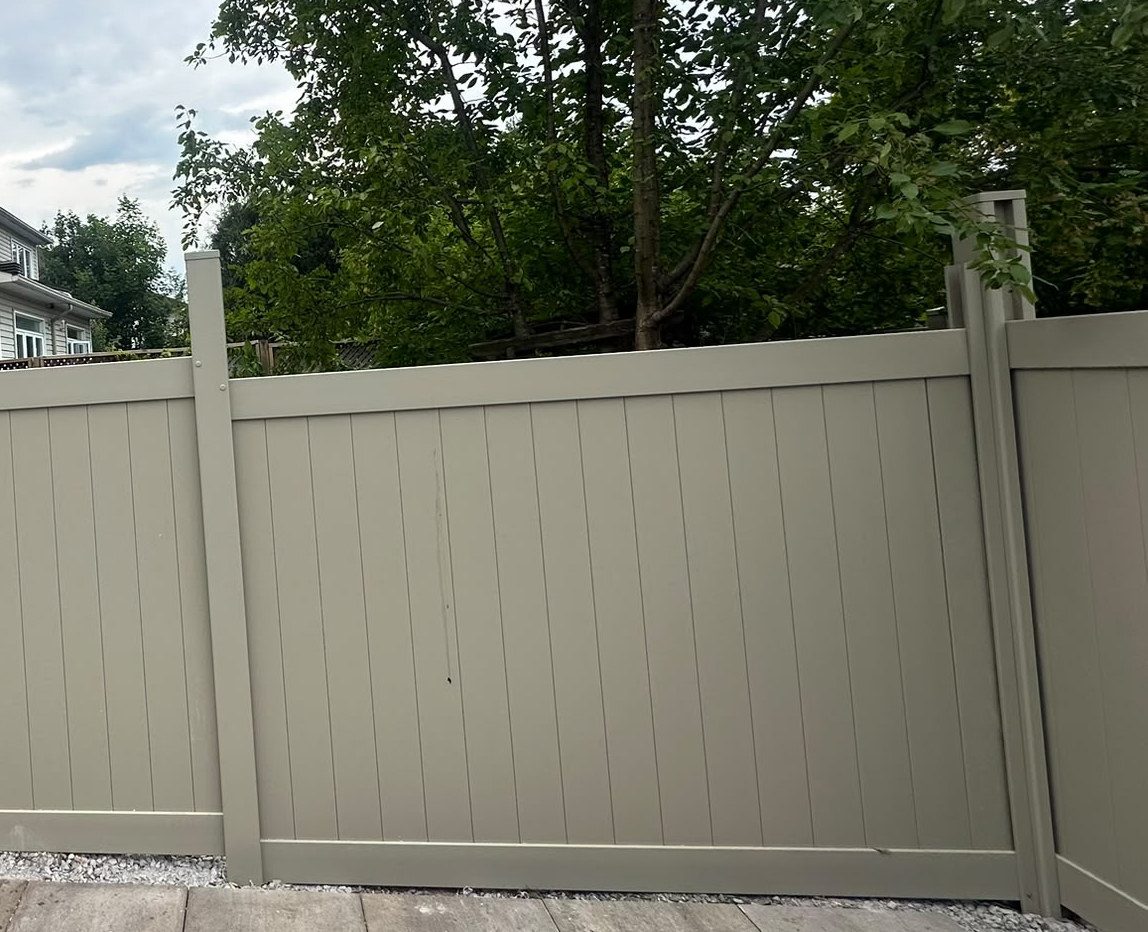 A finished Adobe PVC privacy fence run along a paved side yard, with mature trees behind it.